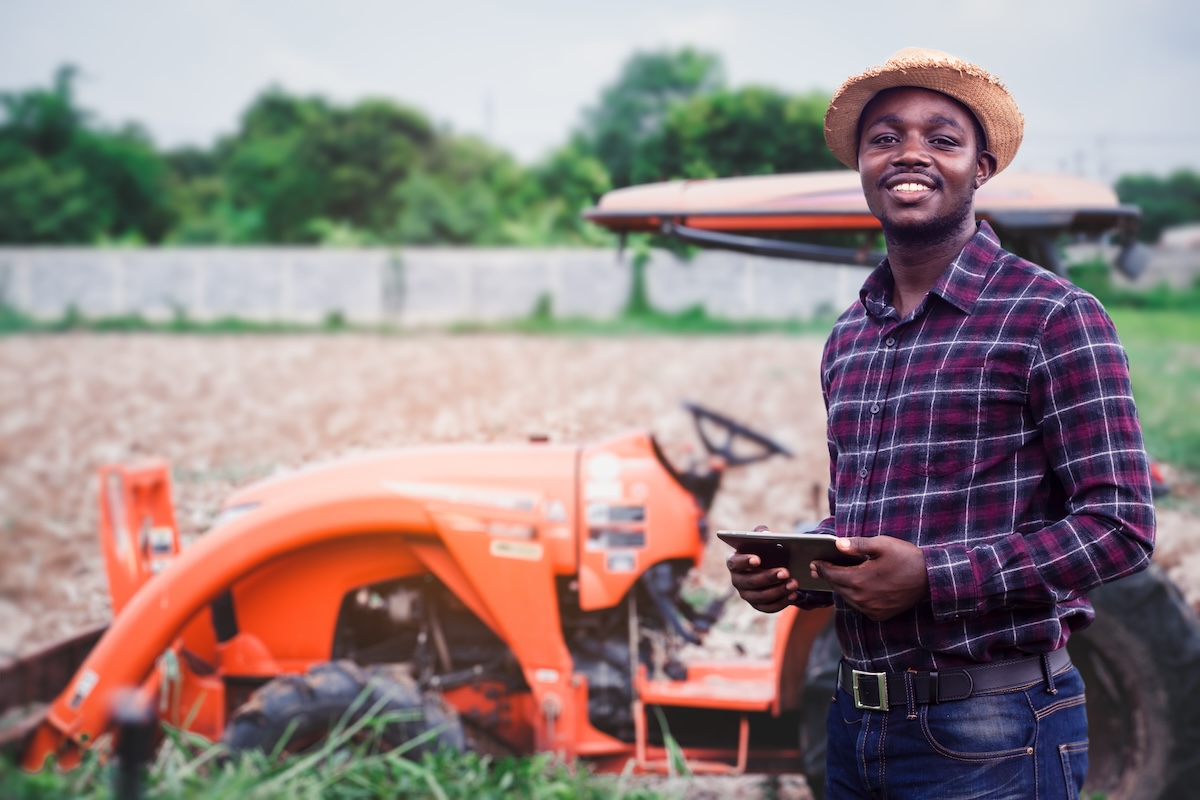 African farmer is using a tablet on the background of working tractor with a cultivator in the field. Agriculture or innovate cultivation concept